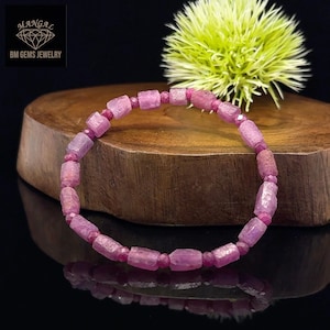 Pode incluir: Uma pulseira de pedras preciosas rosa com contas retangulares e pequenas redondas. A pulseira é exibida em uma superfície de madeira com uma flor verde ao fundo. Joias da BM Gems Jewelry.