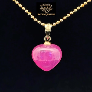 May include: A gold-colored chain necklace with a heart-shaped, pink pendant. The pendant is a vibrant, translucent pink. The necklace has a ball chain design. The pendant is suspended from a gold-colored bail. The background is black.