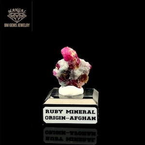 May include: A vibrant ruby mineral specimen from Afghanistan, displayed on a clear stand. The ruby features a rich, deep red hue with white and brown inclusions. The base of the stand has the text "RUBY MINERAL ORIGIN-AFGHAN".