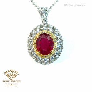 May include: A pendant necklace featuring a central oval-cut red ruby surrounded by a yellow gold halo and two outer layers of clear round-cut stones set in silver-toned metal. The pendant hangs from a silver-toned ball chain necklace.