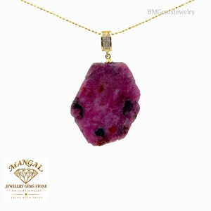 May include: A gold chain necklace featuring a large, raw, irregular-shaped magenta ruby pendant with dark inclusions. The pendant is suspended from a gold bail encrusted with small clear stones. The text BMGemsJewelry appears in the top right corner.