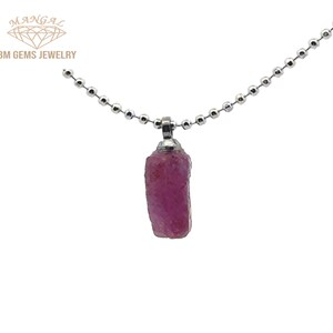 May include: A raw, pink ruby pendant hangs from a silver ball chain. The ruby is a rough, rectangular shape with a textured surface. The jewelry is set against a white background. The brand name "BM GEMS JEWELRY" is visible in the upper left corner.