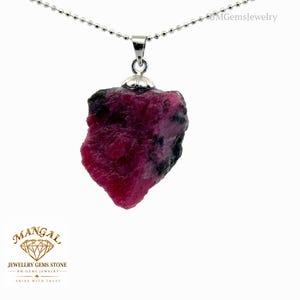 Può includere: Un ciondolo in pietra grezza rosa scuro e nera sospeso a una catena a palline color argento. La pietra ha una superficie irregolare e testurizzata ed è fissata da un cappuccio e un gancio in metallo color argento. Il testo BMGemsJewelry appare in alto a destra.