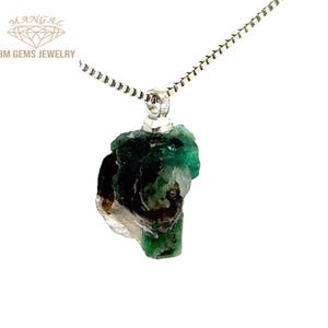 Può includere: Una collana d'argento con un pendente di smeraldo grezzo. Lo smeraldo è di un verde intenso con alcune inclusioni scure e una consistenza naturale ruvida. La collana è esposta su uno sfondo bianco. Il nome del marchio "BM GEMS JEWELRY" è visibile.