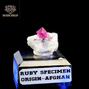 以下が含まれることがあります： アフガニスタン産の鮮やかなピンク色のルビー標本。白い鉱物ベースの上に置かれています。結晶構造を持つルビーは、透明で反射性の高いスタンドに展示されています。「RUBY SPECIMEN ORIGIN-AFGHAN」の文字がスタンドに表示されています。