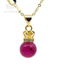 Op de afbeelding: Een gouden ketting met een roze edelsteen hanger. De hanger is bolvormig en heeft een kroon bovenop. De kroon is versierd met kleine, heldere stenen.