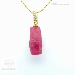 Puede incluir: Un collar de oro con un colgante con una gema de rubí rosa en bruto. El colgante está unido a una cadena de oro con una pequeña fianza con diamantes. El collar se muestra sobre un fondo blanco.