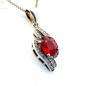 Pode incluir: Um colar de prata com um pingente de rubi oval grande cercado por pedras brancas menores. O pingente está preso a uma corrente de prata.