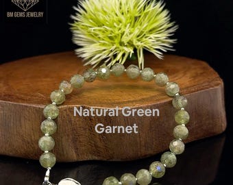 Puede incluir: Un brazalete hecho de cuentas facetadas de granate verde natural. Las cuentas están ensartadas con un cierre plateado. El brazalete se exhibe sobre una superficie de madera con un acento floral verde. El texto "Natural Green Garnet" es visible.