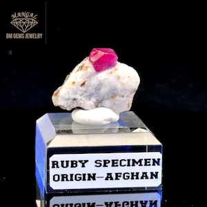 Può includere: Un vivace esemplare di rubino rosso su una roccia bianca e beige, esposto su un supporto rettangolare trasparente. Il supporto presenta un'etichetta con la scritta "RUBY SPECIMEN ORIGIN-AFGHAN". Il logo BM Gems Jewelry è in alto a sinistra.
