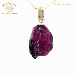 Puede incluir: Un collar de oro con un colgante de rubí en bruto e irregular en tonos magenta intenso y negro. El colgante está unido a un pasador dorado incrustado con pequeñas piedras transparentes. El texto MANGAL BM GEMS JEWELRY SHINE WITH TRUST es visible.