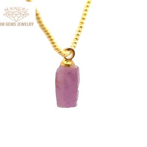May include: A gold-colored chain necklace with a pendant featuring a rough-cut, light purple gemstone. The pendant is attached to the chain with a gold-colored bail. The necklace is set against a white background. The brand name "BM GEMS JEWELRY" is visible.