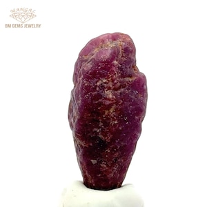 May include: A raw, uncut ruby gemstone with a deep red colour. The natural stone has a textured surface and irregular shape, showcasing its raw, unpolished state. The gemstone is set against a white background, highlighting its colour and form. The image includes the text "BM GEMS JEWELRY".