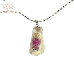 May include: A pendant necklace featuring a raw, uncut gemstone with a vibrant pink ruby embedded in a light-colored matrix. The pendant is suspended from a silver-colored ball chain. The brand name "BM GEMS JEWELRY" is visible.