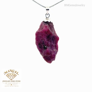 Può includere: Un ciondolo in rubino grezzo color magenta intenso sospeso a una catenina argentata. La pietra preziosa presenta una consistenza ruvida e irregolare con inclusioni scure ed è fissata da un cappuccio in metallo argentato. Il testo BMGemsJewelry appare.