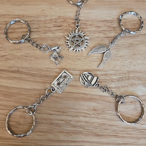 May include: Five silver keychains with different charms. The charms include a cassette tape, a winged angel, a moose, a sun with a pentagram inside, and a small, white, and red striped hat.