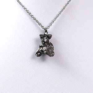 May include: A silver chain necklace with a pendant made of a rough, dark gray, metallic material. The pendant is shaped like a small, irregular rock.