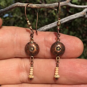 May include: A pair of copper and bone dangle earrings with a textured round copper bead and a small bone bead dangle. The earrings have a hook closure.