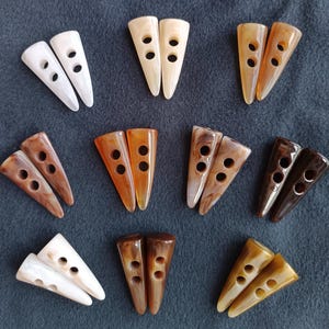 May include: A selection of triangular buttons in shades of brown, cream, and white. Each button has two pairs of holes for sewing. The buttons are displayed on a dark grey surface.