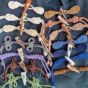 May include: A collection of toggle clasps in various colors, including cream, brown, gray, purple, blue, and green. Each clasp features a horn-shaped toggle and a braided or knotted loop. The clasps are arranged on a dark teal fabric.