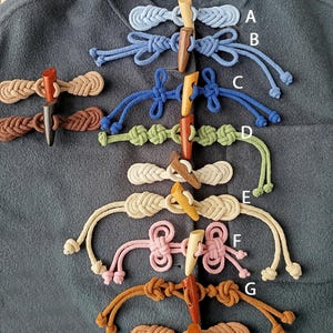 May include: A collection of handmade toggle closures in various colors, including blue, brown, pink, and beige. Each closure features a wooden toggle and braided or knotted rope details. The closures are displayed on a gray fabric background.