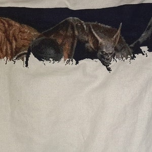 Puede incluir: Una camiseta blanca con un dise&ntilde;o bordado en negro, azul y marr&oacute;n de tres criaturas. Las criaturas son un monstruo marr&oacute;n, un murci&eacute;lago y una figura encapuchada.