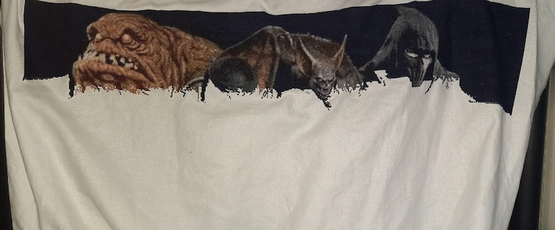 Puede incluir: Una camiseta blanca con un dise&ntilde;o bordado en negro, azul y marr&oacute;n de tres criaturas. Las criaturas son un monstruo marr&oacute;n, un murci&eacute;lago y una figura encapuchada.