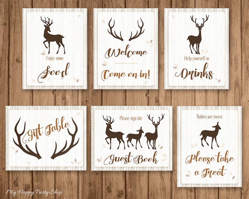 May include: Set of six decorative signs with a rustic theme. Each sign features a brown deer silhouette or antlers on a cream background with text like "Welcome" and "Drinks". The signs are arranged on a wooden surface.