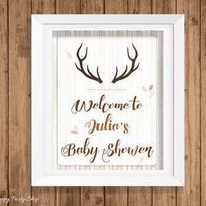 May include: A framed sign with a white background and brown text that reads "Welcome to Julia's Baby Shower." The sign features brown antlers at the top and is decorated with small leaf accents. The frame is white, and the background is a wood grain.