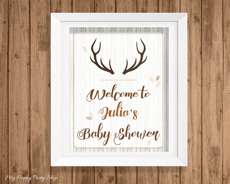 May include: A framed sign with a white background and brown text that reads "Welcome to Julia's Baby Shower." The sign features brown antlers at the top and is decorated with small leaf accents. The frame is white, and the background is a wood grain.