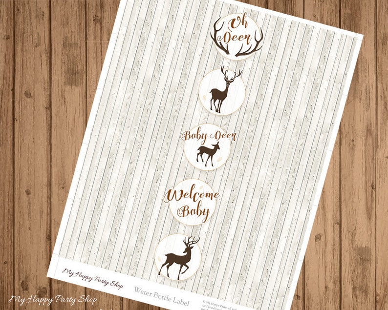 May include: Water bottle labels with a woodland theme. The labels are circular with a light beige background and brown text and deer silhouettes. Text includes "Oh Deer", "Baby Deer", and "Welcome Baby". The labels are on a sheet with a light wood grain background.