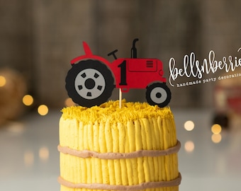 May include: A red and black tractor cake topper on a yellow and brown cake. The cake is decorated to look like a bale of hay. The cake is on a wooden cake stand. The background is a rustic wooden wall with hay bales and string lights.
