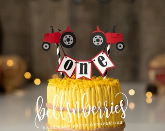 May include: A yellow and brown cake decorated with red toy tractors and a banner that says "one". The cake is on a wooden cake stand. The background is a rustic barn scene with hay bales and string lights.