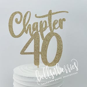 May include: A white cake with a gold glitter cake topper that reads "Chapter 40". The cake is on a white cake stand. The cake topper is made of gold glitter cardstock. The cake has a textured, ridged design.