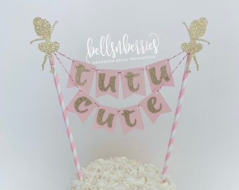 May include: A white frosted cake with a pink and gold glitter banner that reads "tutu cute". The banner is held up by pink and white striped straws, and topped with gold glitter fairy cutouts. The cake is decorated with white frosting roses.