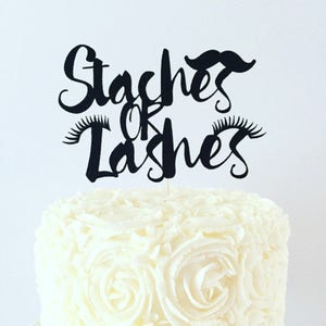 May include: Black cake topper with the text "Staches or Lashes" in a script font. The topper features a mustache and eyelashes.