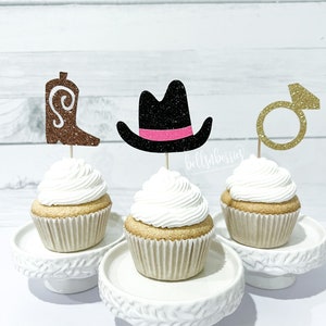 May include: Three cupcakes decorated with glitter toppers. The toppers are a brown cowboy boot, a black cowboy hat with a pink band, and a gold ring with a diamond. The cupcakes are on white cake stands.