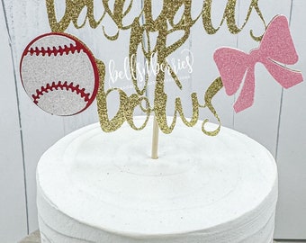 May include: Gold glitter cake topper with the text "baseballs & bows" and a pink bow and a red and white baseball.