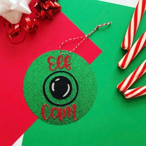 May include: Green glitter Christmas ornament with the text "Elf Cam" and a black and white eye graphic.