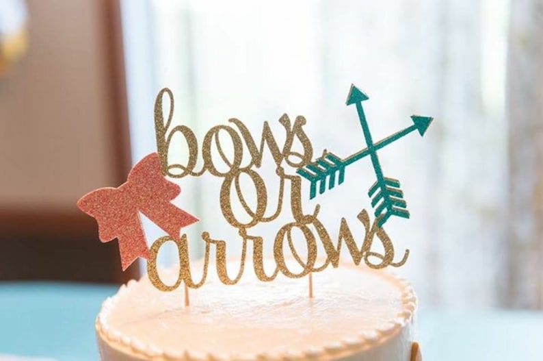 May include: Gold glitter cake topper with the words "bows or arrows" and a pink glitter bow on the left and two teal glitter arrows on the right.