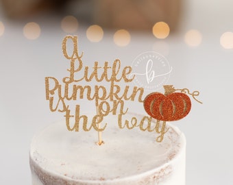 Könnte beinhalten: Ein goldener Glitzer-Kuchentopper mit dem Text "A Little Pumpkin is on the way" und einem kleinen orangefarbenen Glitzerkürbis.