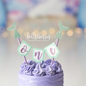 May include: Purple frosted cake with a green and purple "one" banner and mermaid tail toppers. The banner says "bellsnberries handmade party decorations".