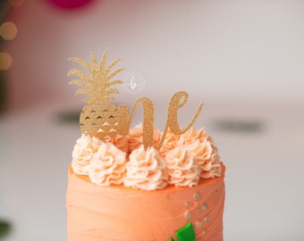 May include: A pink and orange cake with gold glitter frosting and a gold glitter pineapple and the word "one" on top.