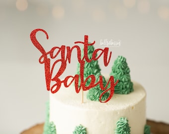 May include: A white frosted cake with green frosting trees and a red glitter "Santa Baby" cake topper. The cake is on a wooden base, with a blurred background.