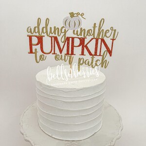 May include: A white cake with red and gold glitter lettering that reads "adding another pumpkin to our patch" with a small white glitter pumpkin on top. The cake is on a white cake stand.
