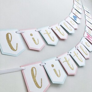 May include: A pink and blue banner with gold glitter letters spelling "little" in a cursive font. The banner is strung together with white ribbon.