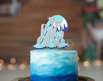 May include: A blue ombre cake topped with a glittery silver and blue cake topper that reads "The Big One". The cake topper is shaped like a wave. The cake is on a silver tray.