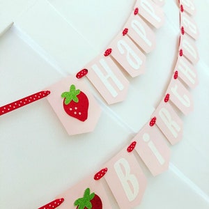 May include: Pink and white banner with the words "Happy Birthday" and red and green strawberry illustrations. The banner is strung with red and white polka dot ribbon.