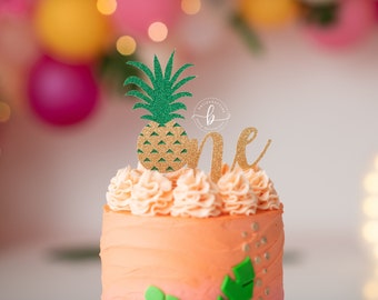 May include: A pink and orange cake with a pineapple and the word "one" on top. The cake is decorated with green leaves and gold glitter.