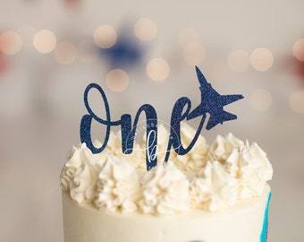 Könnte beinhalten: Ein weißer Kuchen mit blauer Zuckerglasur und einem blauen Glitzer-Kuchentopper, auf dem "one" mit einem Flugzeug steht.